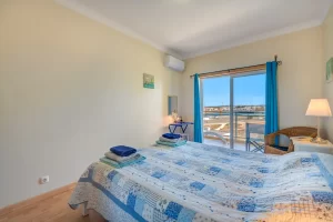A bright bedroom featuring a double bed with a blue and white patchwork quilt and folded blue towels. On the right, a white nightstand holds a lit table lamp next to a wicker armchair. Large sliding glass doors with vibrant turquoise curtains open onto a sunny balcony, offering an open view of a coastal landscape under a clear blue sky. An air conditioning unit is mounted high on the wall.