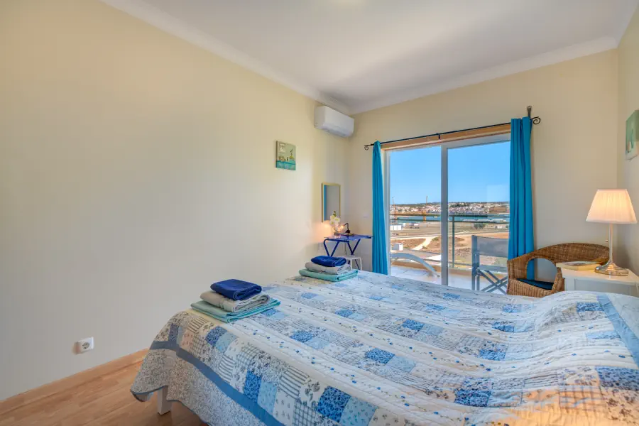 A bright bedroom featuring a double bed with a blue and white patchwork quilt and folded blue towels. On the right, a white nightstand holds a lit table lamp next to a wicker armchair. Large sliding glass doors with vibrant turquoise curtains open onto a sunny balcony, offering an open view of a coastal landscape under a clear blue sky. An air conditioning unit is mounted high on the wall.