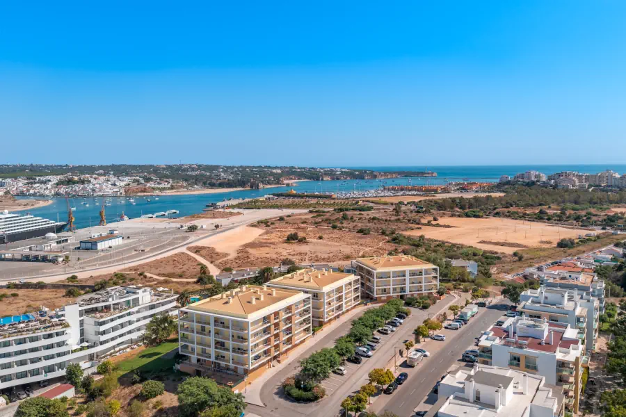 A high-angle aerial panorama showcasing several modern apartment complexes with tiled roofs along a paved street with parked cars. Beyond the buildings, a wide blue river estuary opens up, featuring a large cruise ship docked on the left port and a marina filled with boats in the center. The vast blue ocean stretches across the horizon under a clear sky.