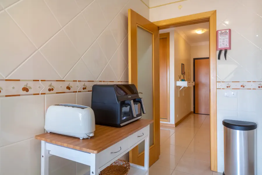 A close-up view of a small white kitchen utility table with a wooden top, holding a white two-slice toaster and a black dual-basket air fryer. The background features light beige tiled walls with a decorative fruit border and an open wooden glass-panel door that leads out to a brightly lit hallway with polished tile flooring.