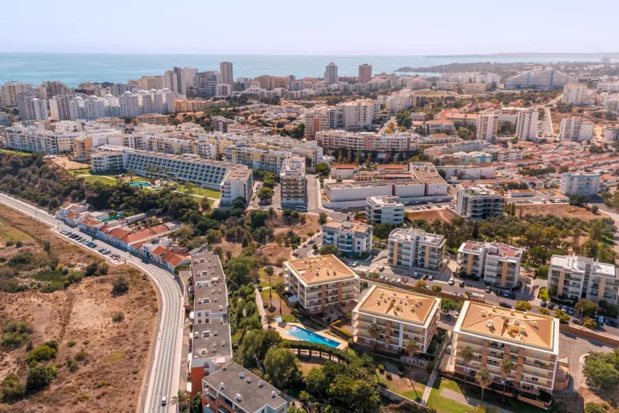 A high-altitude aerial view overlooking a dense coastal town filled with numerous multi-story apartment complexes and residential streets. In the lower center, the three distinct yellow-roofed buildings of the complex with its private swimming pool are clearly visible. A winding road borders a dry, open field on the left, while the vast blue ocean stretches along the horizon in the background.