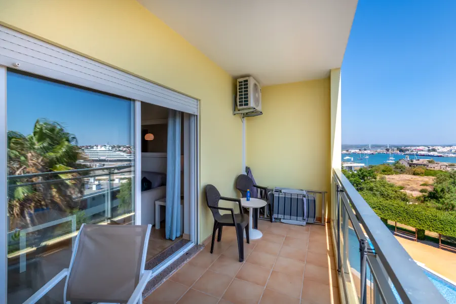 A long tiled balcony with light yellow walls and a glass railing, featuring two grey plastic chairs and a small white round table in the corner. An air conditioning unit is mounted high on the wall above the seating. Large sliding glass doors lead into the apartment, reflecting a palm tree, while the balcony offers wide views of the river and distant town to the right.