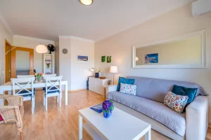 A bright living and dining room with light wood floors. In the right foreground sits a grey sofa adorned with patterned and dark teal cushions, positioned beneath a large white-framed horizontal mirror. To the left, a white dining table is surrounded by four white chairs with blue seat cushions, illuminated by an arched floor lamp. The background shows white storage cabinets and framed artwork on the walls.