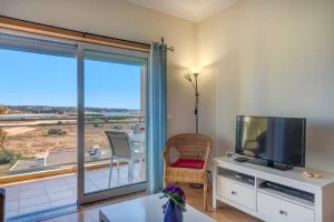 An indoor view of a living space next to large sliding glass doors with light blue curtains that open onto a sunny balcony. On the right, a television sits on a white media console next to a wicker armchair with a red cushion and a tall floor lamp. Through the glass doors, a balcony with a white table and chairs is visible, overlooking a wide, dry landscape and a distant coastline.