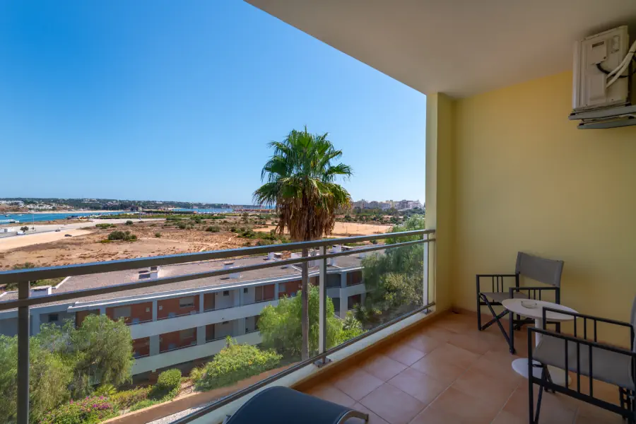 A view from a tiled balcony with a metal railing, featuring outdoor seating with two chairs and a small round table on the right. A tall palm tree stands directly in front of the balcony, overlooking a lower building and a vast, dry coastal landscape with a harbor and town visible in the distance under a clear blue sky.