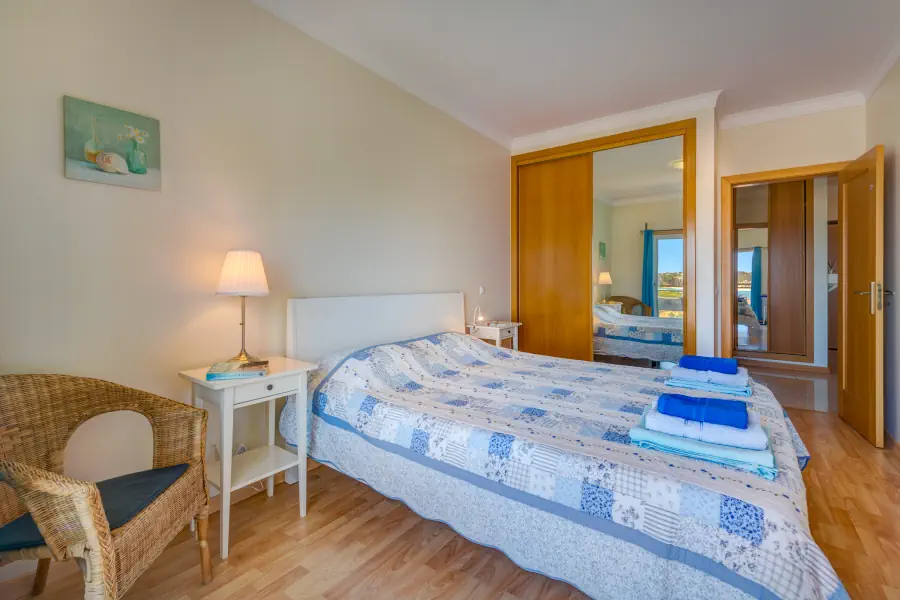 A second angle of the bedroom showing a double bed with a blue and white patchwork quilt and stacked towels. To the right is a large wooden built-in wardrobe with a full-length mirrored sliding door that reflects the balcony window. The foreground features a woven wicker armchair and a white nightstand with a lit table lamp.