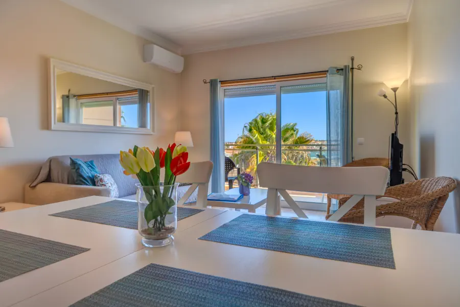 Een licht overgoten woon- en eetkamer met een glazen schuifdeur die uitkijkt op een zonnig balkon met een palmboom. Op de voorgrond staat een witte eettafel met blauwe placemats en een glazen vaas met rode en gele tulpen. Op de achtergrond zijn een bank, een spiegel en rieten stoelen zichtbaar.