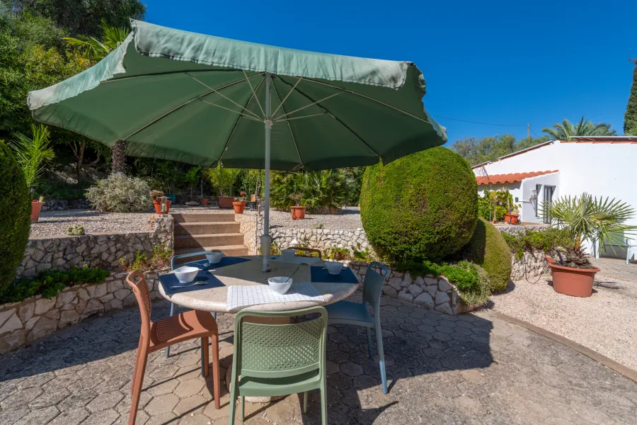 Une terrasse pavée ensoleillée avec une table ronde dressée sous un grand parasol vert, entourée de plantes méditerranéennes.