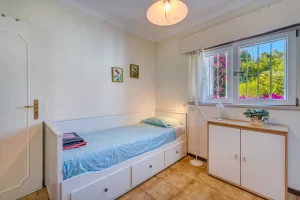 Une chambre lumineuse avec un lit simple blanc muni de tiroirs, une commode, deux petits tableaux et une fenêtre donnant sur des bougainvilliers.