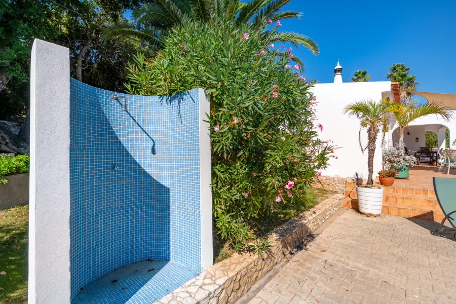 Une douche extérieure courbée avec de la mosaïque bleue, située dans un jardin ensoleillé à côté d'une villa blanche.