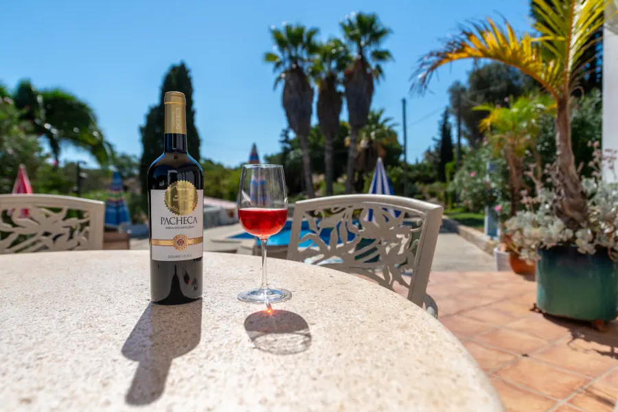 Une bouteille de vin Pacheca et un verre de vin rosé posés sur une table de terrasse ensoleillée près d'une piscine.