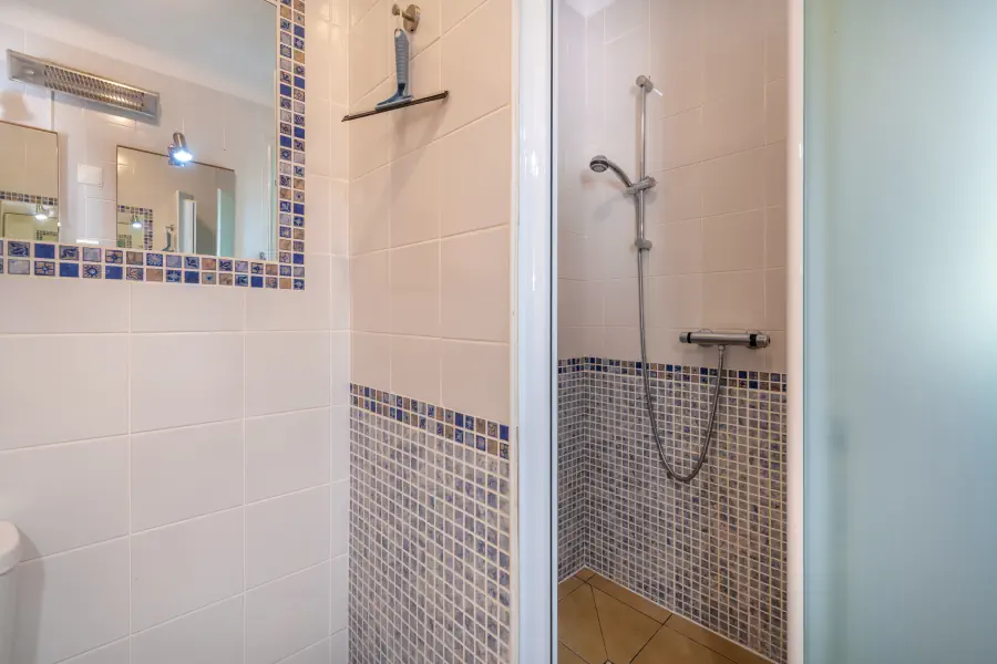 Une cabine de douche carrelée de mosaïque bleue et grise, à côté d'un mur en carrelage blanc avec un miroir